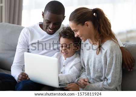Cheerful mixed race family sitting together on comfortable sofa in living room, holding laptop on knees, looking at screen, playing online game, watching movie or cartoons, shopping in internet store.