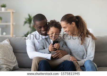 Smiling young woman sitting at living room on comfortable couch near happy african american husband holding cute small mixed race boy on knees, watching little kid son playing on mobile phone. Royalty-Free Stock Photo #1575811945