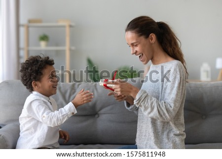 Astonished excited young woman getting wrapped gift package from happy mixed race little cute son, sitting on comfortable sofa in living room at home, Mothers Day or birthday celebration concept.