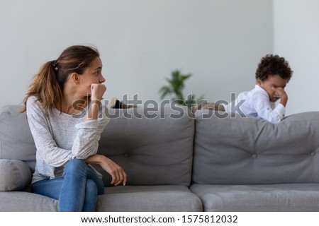 Unhappy young woman sitting on sofa in living room at home, looking at stressed offended punished upset little kid son, lying on couch. Sad mommy feeling guilty about quarrel conflict with child. Royalty-Free Stock Photo #1575812032