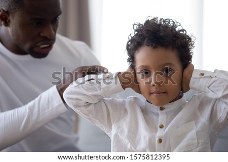 Head shot portrait little mixed race preschool boy covering ears with hands, looking at camera, don t want listen shouting or blaming african american angry father. Family relations problem concept. Royalty-Free Stock Photo #1575812395