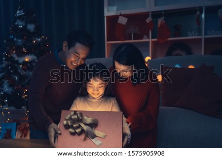 Happy family and little girl open magic gift box with light shining and kiss in living room that decorated with Christmas tree for Christmas festival coming soon, Asian Christmas family concept