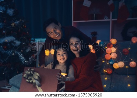 Happy family and little girl open magic gift box with light shining and kiss in living room that decorated with Christmas tree for Christmas festival coming soon, Asian Christmas family concept