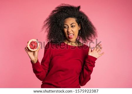 African american girl act ape her face from grapefruit cid. Woman grimaces. Lady with curly hairstyle on pink background. Diet. healthy lifestyle concept.