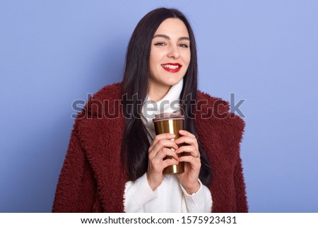 Picture of beautiful young brunette woman wearing winter jacket and holding thermo mug with coffee or tea in studio, isolated over blue background, having happy facial expression. People concept.