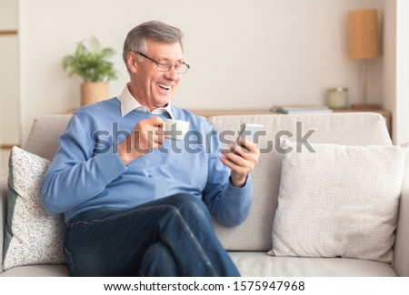 Happy Senior Gentleman Using Smartphone Texting And Having Coffee Sitting On Sofa At Home. Selective Focus Royalty-Free Stock Photo #1575947968