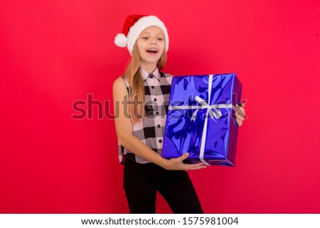 Cute little girl wearing Christmas hat standing isolated over red background, holding blue present box