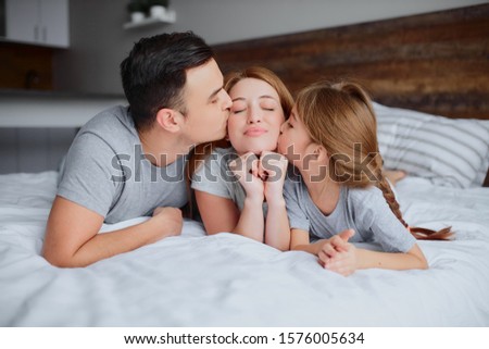 portrait of young caucasian family lying together on bed, child girl and father kissing mother, young parents and child girl, happy together at home, spend free time at home on bed