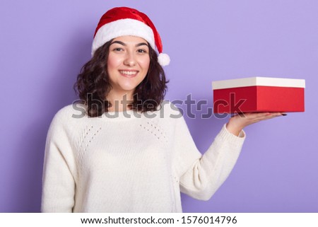 Image of smiling good looking young girl holding red and white box on palm hand, looking directly at camera, preparing for new year, wearing santa claus hat and white sweater. Present concept.