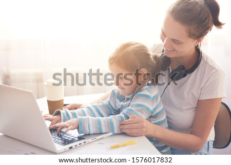 Happy family concept. Front view of adorable little girl sitting on mother laps while watching cartoons online, mother and her child spending time together, mom and kid wearing casual outfits.