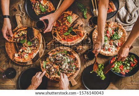Family or friends having pizza party dinner. Flat-lay of people cutting and eating Italian pizza and drinking red wine from glasses over wooden table, top view. Fast food lunch, gathering, celebration