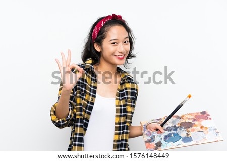 Young artist woman holding a palette over isolated white background showing ok sign with fingers