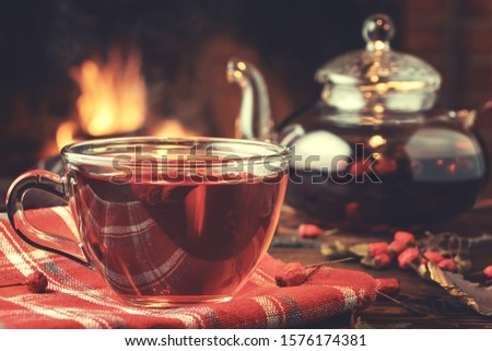 Tea with hawthorn in a glass cup and teapot on a wooden table in a room with a burning fireplace, closeup