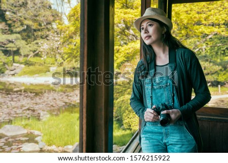 elegant lady photographer travel in osaka japan. beautiful woman tourist holding camera while leaning against window by spring pond in japanese park. female backpacker watching view of lake in garden