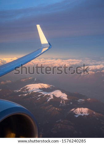 Flying over the Alps during the sunset in winter time. Aerial view from the airplane window. Mountains and peaks covered by fresh snow. A wing and engine of the airplane