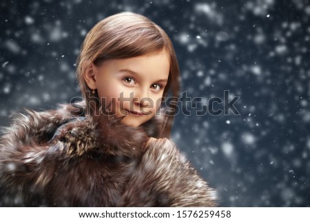 Christmas winter kids fashion. Cute little girl in a silver fox fur coat stands outdoor under the snowfall and smiles. Evening, Santa Claus house in the background. Copy space.