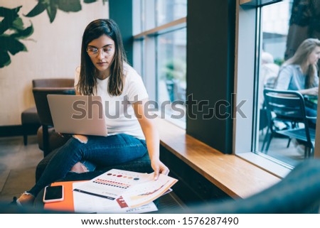 Young female designer in casual clothes and eyeglasses using laptop and color palette while working in lounge zone and drawing new project 