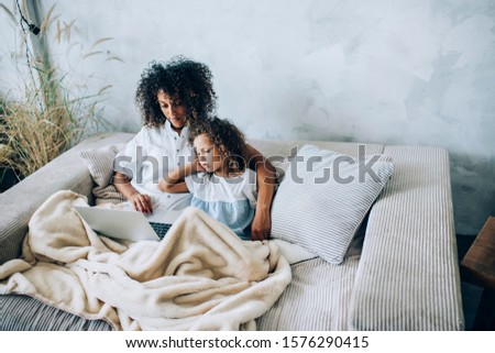 From above concentrated young ethnic mother cuddling cute little daughter and using laptop while resting together on soft sofa with blanket in light flat