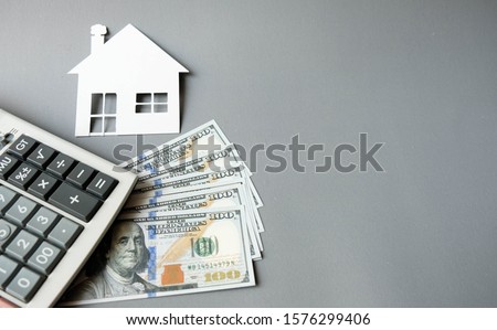 White paper house with dollars banknotes and calculator on a grey background. Concept of save, investment in real estate, refinance, retention, collect money for buy, sell, decorate or repair for home Royalty-Free Stock Photo #1576299406