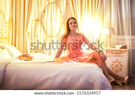 Girl in pink dress with present in the box on the bed with white linen. Model posing during fashion photoshoot