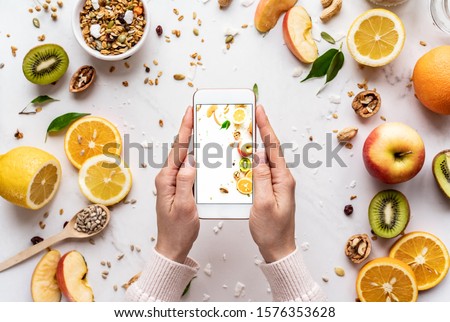 Female hands holding smartphone take photo on healthy food background, woman using phone mobile apps make digital picture on screen of diet nutrition vegan fruit granola seed on white table, top view