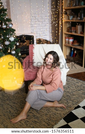 The concept of Christmas home shooting. Beautiful attractive cute brunette girl with short hair in warm home clothes sitting on a chair near the Christmas tree on the background of the fireplace.