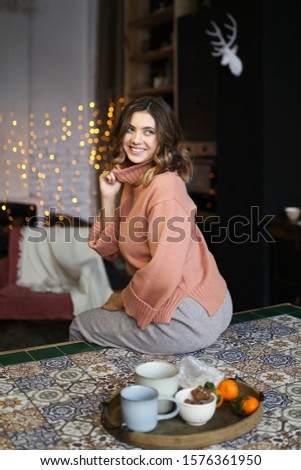 The concept of Christmas home shooting. Beautiful attractive cute brunette curly girl in warm home clothes laughs and poses against the background of Christmas dark kitchen, garlands and bokeh.