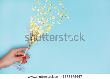 Woman hand holding martini glass with pouring out golden stars confetti on blue background. Top view. Flat lay.