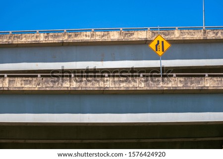 Warning traffic road sign indicating a narrowing of the road is located on one of the levels of a multi-level road overpass against a blue sky