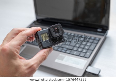 Action camera in hand. Laptop in background. Concept of transferring and storing data from a camera to a computer.
