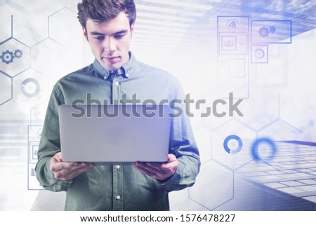 Serious young engineer in casual clothes looking at laptop in modern city with double exposure of blurry business interface. Concept of technology. Toned image