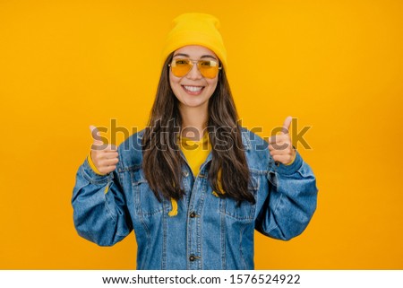 Happy stylish young woman in yellow hat and sunglasses showing thumbs up on yellow background