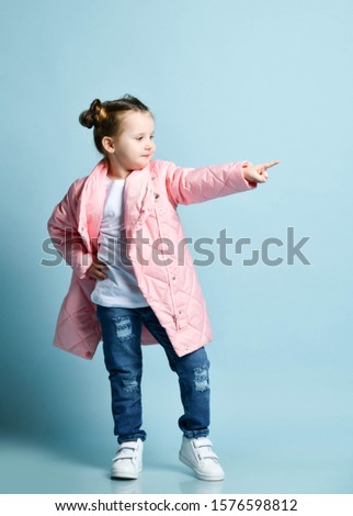 Stylish happy frolic little kid girl with funny buns and in winter jacket white t-shirt, modern blue torn jeans is pointing her finger aside