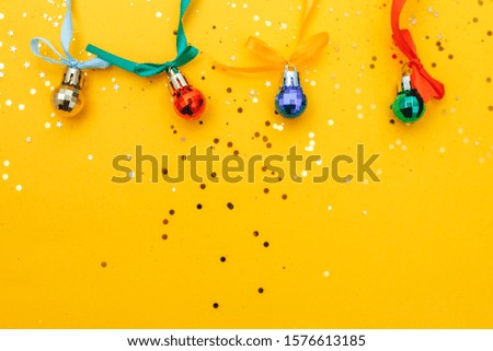 Color christmas balls with different ribbons and bows hanging on yellow background with gold sparkles, flat lay, top view.
