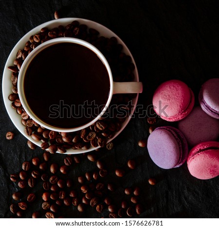 A white cup with black fragrant coffee stands on a black background with coffee zurnes next to colorful french cookies macaroons