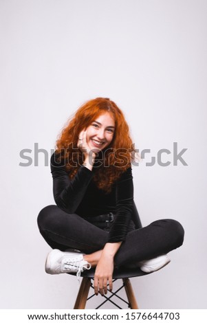 Lovely young girl is sitting on the chair and smiling near white wallpaper.