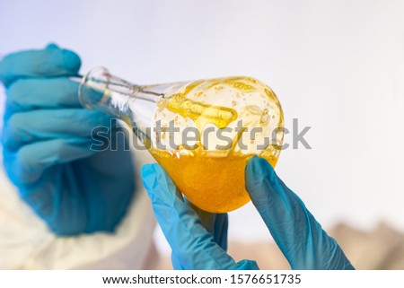 A test tube with a yellow viscous liquid in the hands of a chemist. Gloved hands holding a flask of yellow liquid chemical composition. Analysis of raw materials for production. Chemical experiment. Royalty-Free Stock Photo #1576651735