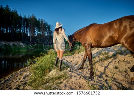 a woman in white dresses and white hats at sunset on a summer evening led the horse to the lake. rural life