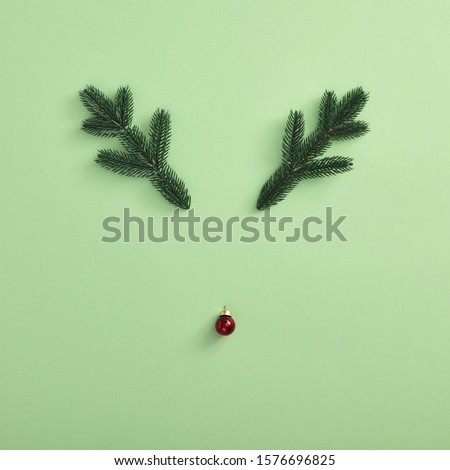 Christmas minimal concept - reindeer face made of red xmas ball and evergreen branch on green background. Square composition, flat lay, top view. Merry christmas holiday. Winter holiday symbol.
