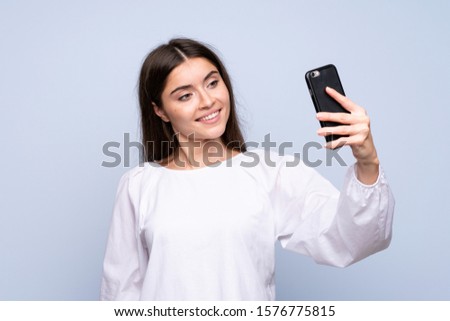 Young woman over isolated blue background taking a selfie with the mobile