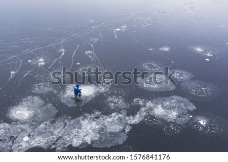 Winter landscape. The frozen surface of the river is covered with ice patterns. On the ice sits a lone fisherman. The view from the top.