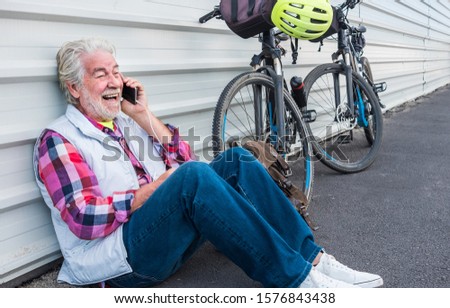Cheerful senior man sitting  against a white metal wall. Listening something fun at his mobile phone. Backpack close to him. Active people with his electric bicycle black color and a yellow helmet