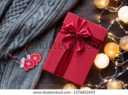 Red gift box with bow on dark wooden background. Festive garland with lights. Cozy picture.
