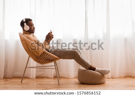 Weekend. Relaxed Black Man In Wireless Headset Using Mobile Phone Listening To Audiobook Sitting On Modern Chair Against Window Indoor