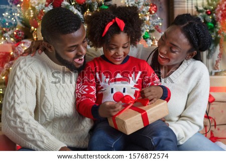 Cute little black girl opening Christmas gift with her parents, sitting together in decorated living room.