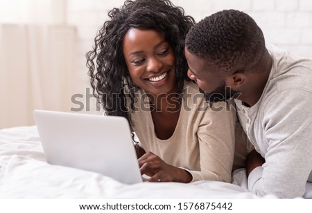 Smiling young afro couple using laptop in bed at home, browsing social media or surfing internet, free space