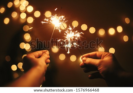 Glowing sparklers in hands on background of golden christmas tree lights, couple celebrating in dark festive room. Happy New Year. Space for text. Fireworks burning in hands. Happy Holidays Royalty-Free Stock Photo #1576966468