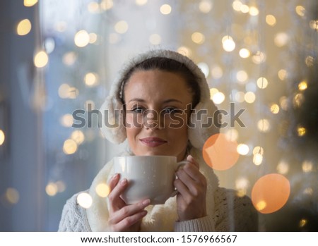 Beautiful girl with earmuffs holding cup of hot coffee with christmas lights in background. Festive moments concept