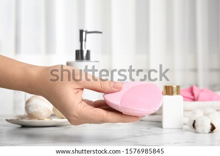 Woman holding face cleansing brush over marble table, closeup. Cosmetic accessory