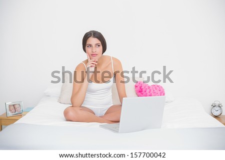 Thinking young brown haired model in white pajamas using a laptop in bright bedroom